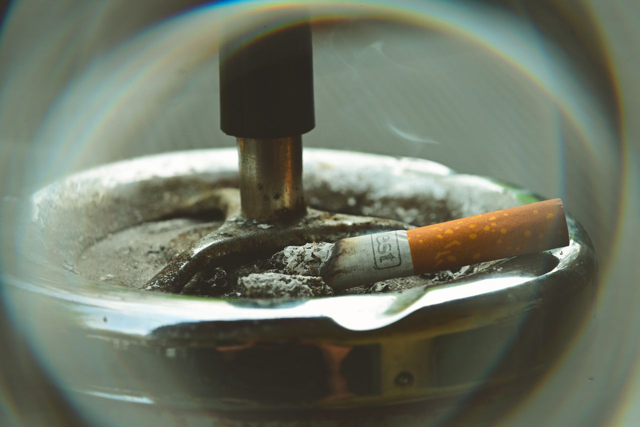 A smoldering cigarette butt resting in an ashtray surrounded by smoke and ash.